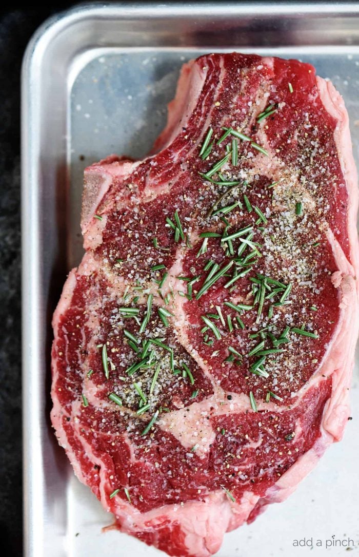 Baking sheet with a raw rib eye steak with generous marbling. It is sprinkled with Stone House seasoning and rosemary // addapinch.com