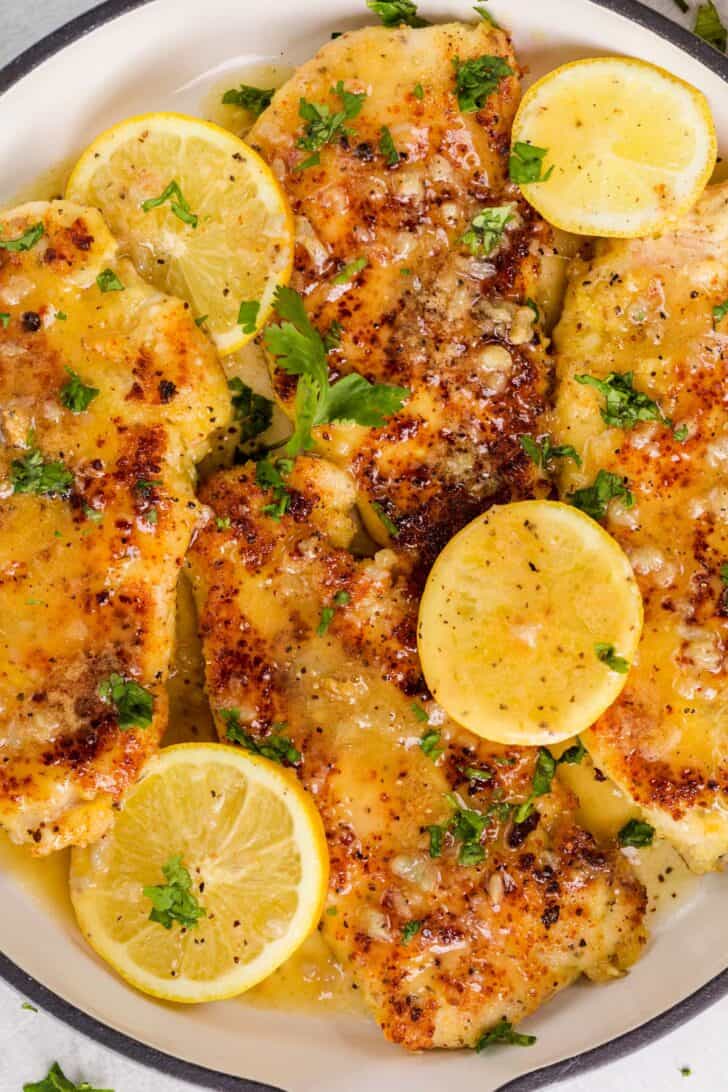 Homemade lemon chicken recipe in a skillet topped with a lemon butter sauce and fresh lemon slices then garnished with herbs.